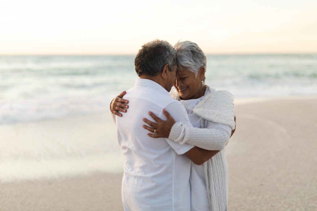 Happy newlywed senior biracial bride and groom hugging each other at beach during sunset